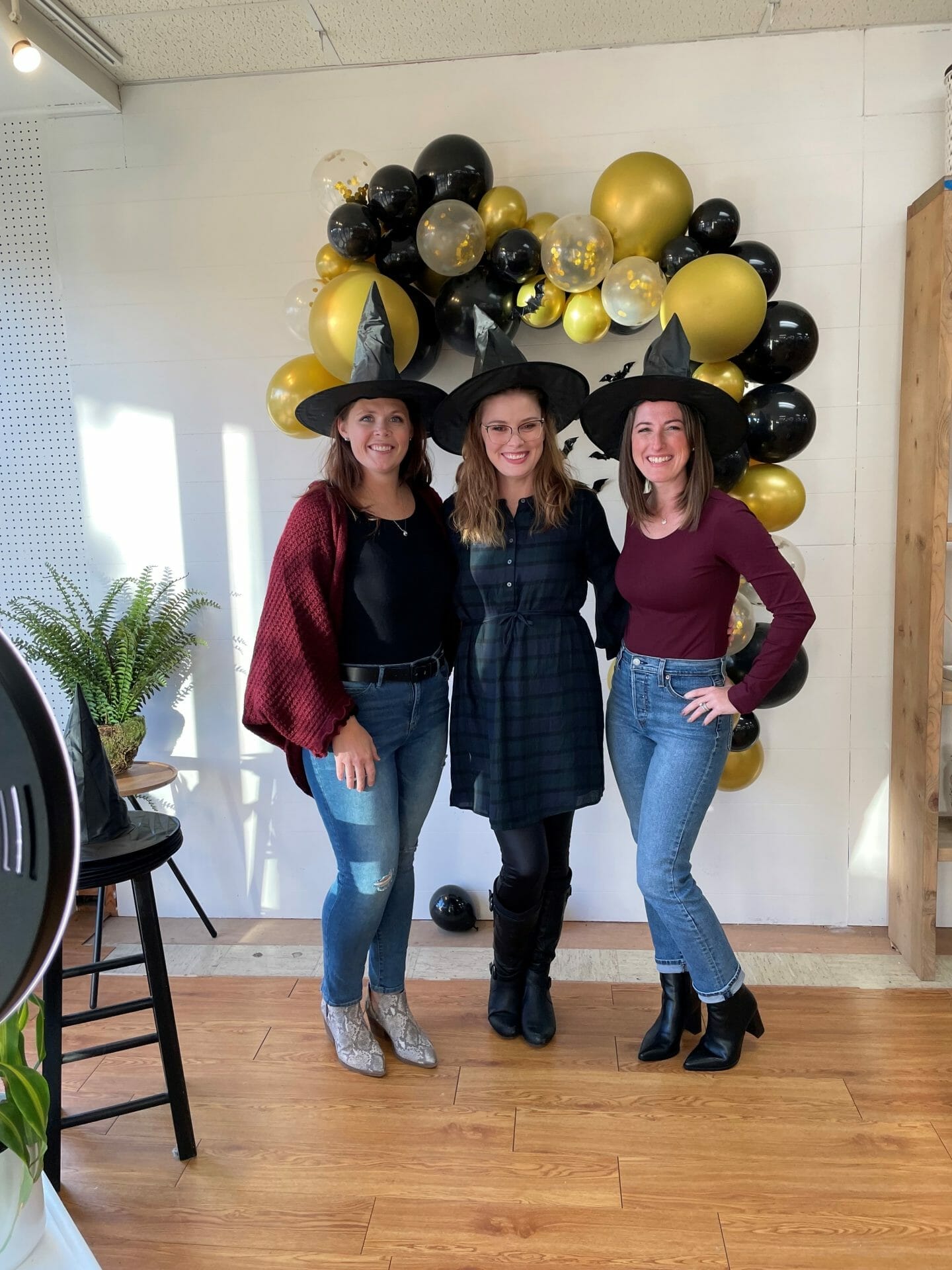 Three ladies in witches hats posed in front of gold and black baloons.